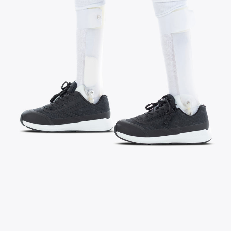 A person with prosthetic legs wears Black BILLY Goat orthotic shoes by BILLY Footwear, featuring white soles, standing on a plain white background.