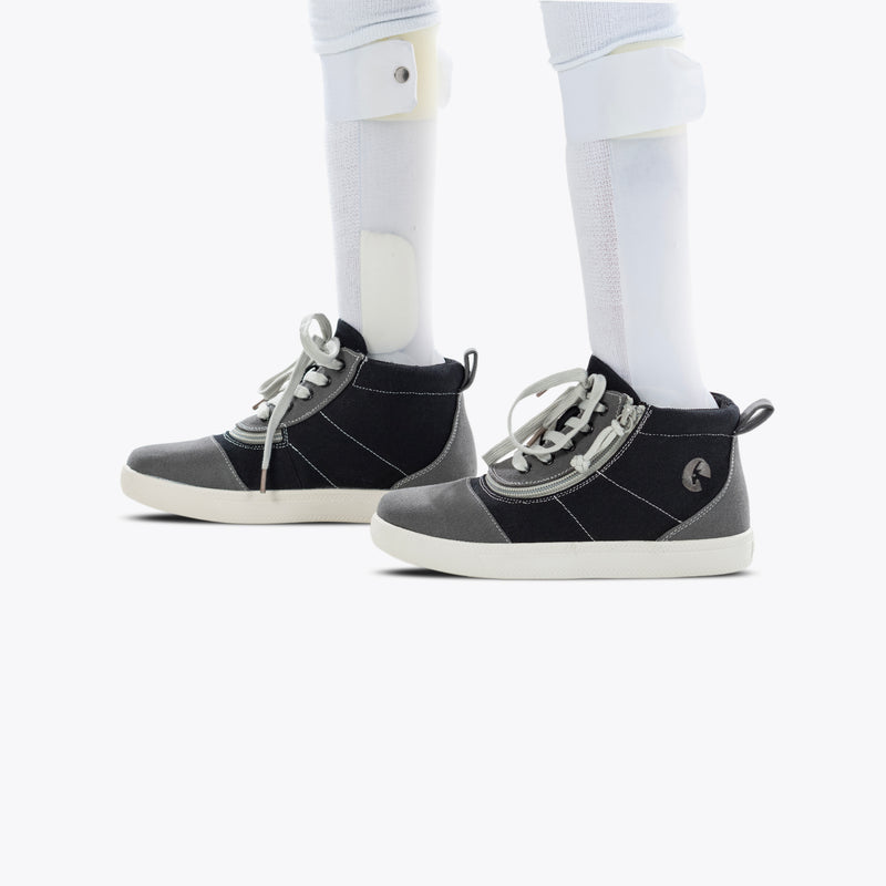 A person stands on a white background, wearing FINAL SALE - Grey Colorblock BILLY D|R Short Wrap High Tops by BILLY Footwear with white soles and orthotic leg braces over white socks.