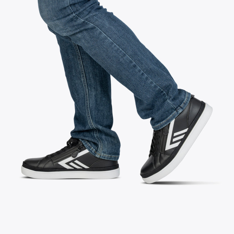 A person in blue jeans and Men's Black/White BILLY CS Low sneakers by BILLY Footwear, with double zippers and white soles, walks against a plain white background.