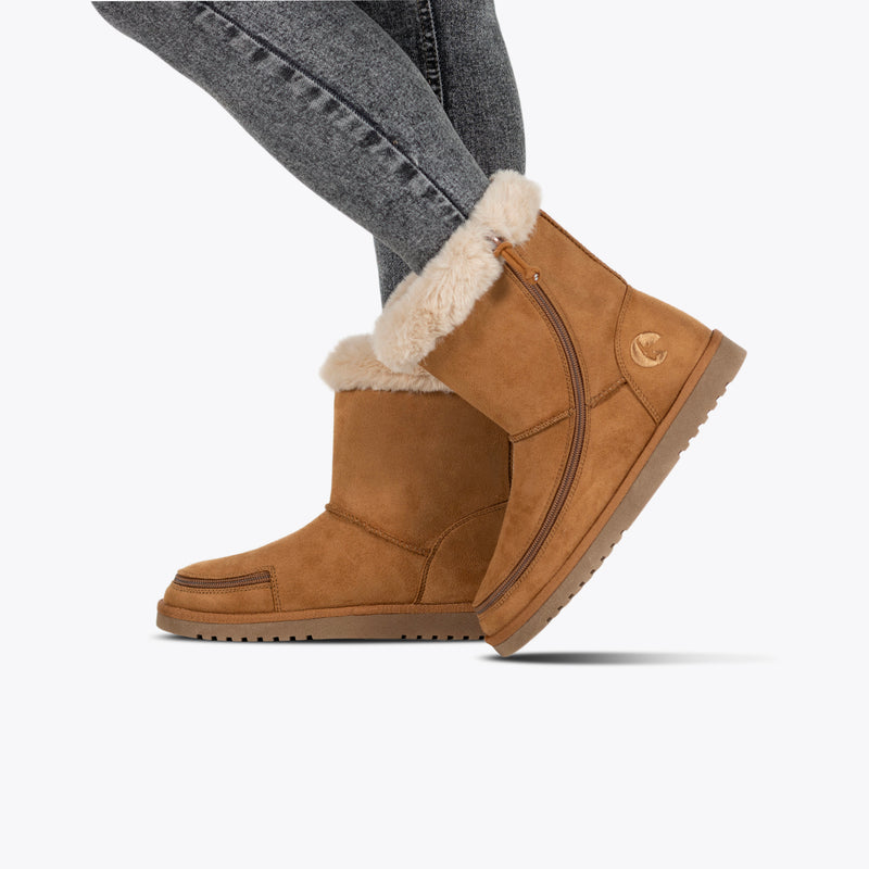 A person wears Women's Chestnut BILLY Cozy II boots by BILLY Footwear with side zippers and gray skinny jeans, standing on a white background.